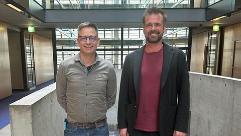 Korrespondent Uwe Berndt und Timo Lange von Lobby Control stehen in der Halle im ARD-Hauptstadtstudio nebeneinander und schauen in die Kamera