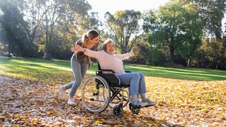 Eine jüngere Frau schiebt eine ältere im Rollstuhl, beide lachen.