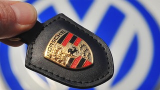 Ein Porsche-Firmenlogo wird vor ein VW-Logo in Wolfsburg gehalten