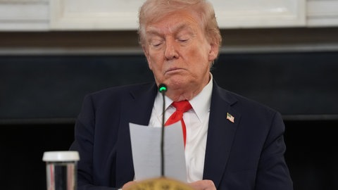 US-Präsident Donald Trump sitzt vor einem Mikrofon und hält einen Zettel in der Hand.