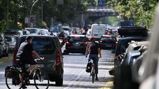 Autos, Fahrradfahrer und Fußgänger sind auf einer Straße unterwegs.