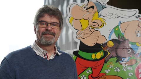 Der Asterix-Übersetzer Klaus Jöken steht vor einer großen Asterix-Zeichnung