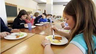 Kinder einer Ganztagsgrundschule sitzen an einer langen Reihe zusammengeschobener Tische und essen Fischstäbchen, Kartoffelbrei und Salat.