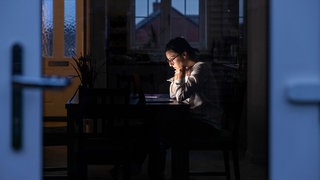 Eine Frau sitzt am Schreibtisch und schaut nachdenklich in ihren Laptop, während es draußen dunkel ist