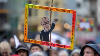 Eine Teilnehmerin der Kundgebung hält eine gemalte und aus Pappe ausgeschnittene Figur von Bundeskanzler Merz hoch.