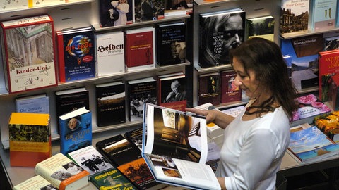 Eine Frau blättert auf den Stuttgarter Buchwochen in einem Buch. Hinter ihr ist ein Regal voller Bücher aufgebaut. Die Frau trägt ihre dunkelbraunen Haare offen. Sie fallen lässig über ihre Schultern auf den Rücken.