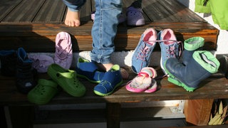 Kinderschuhe in verschiedenen Größen stehen auf einer Treppe, während ein Kind in Jeans und ohne Schuhe über die Schuhe auf der mittleren Stufe steigt.