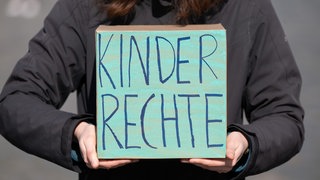 Eine Frau hält einen Karton mit dem Schriftzug „Kinderrechte“. Sie trägt einen grauen Pullover. Ihr Gesicht sieht man nicht.