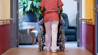 Beliebteste Pflegerin: Eine Mitarbeiterin in der Pflege schiebt eine Person in einem Rollstuhl über eine Krankenhaus-Flur.