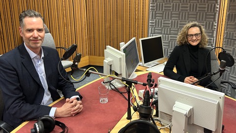 Christian Dürr und Lissy Kaufmann sitzen in einem Tonstudio an einem Tisch mit Mikrofonen und Rechnern und schauen in die Kamera
