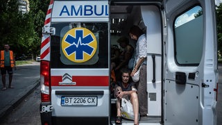 Verletzte Einwohner in einem Krankenwagen nach einem russischen Artillerieangriff in Cherson. Eine Tür des Wagens ist geöffnet. Man sieht eine verletzte Person, die auf dem Boden sitzt. Daneben steht eine weitere.