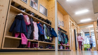 Kinder rennen in einer Kindertagesstätte einen Gang mit vielen Jacken und Schuhen entlang.