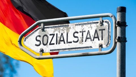 Ein altes Schild "Sozialstaat" vor deutscher Flagge symbolisiert den Druck auf das deutsche Sozialsystem.