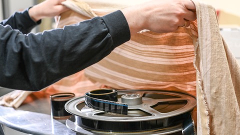 Ein Mitarbeiter deckt in einem Kino-Vorführraum alte Filmrollen ab, die unter einem beige gestreiften Tuch lagen.