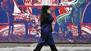 Eine Frau, die ein Hidschab Kopftuch trägt, geht telefonierend an einem Wandbild in Teheran vorbei, das Iran und die Vereinigten Staaten am Verhandlungstisch zeigt.