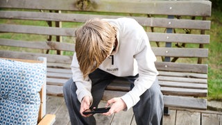 Ein Jugendlicher sitzt auf einer Bank und schaut auf sein Smartphone. Er ist vornübergebeugt, seine blonden Haare fallen über sein Gesicht.