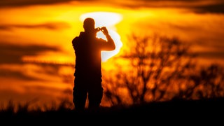Ein Mann fotografiert die Sonne, die farbenprächtig am Rand eines Feldes untergeht.