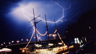 Die Aufnahme zeigt ein Gewitter über der Innenstadt von Mainz. Im Hintergrund die Theodor-Heuss-Brücke, vorne eine mit Lampen geschmückte Dreimastbark.