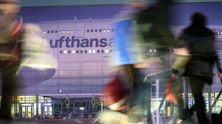 Eine Projektion mit dem Logo der Lufthansa ist auf der Außenwand eines Gebäudes am Flughafen Frankfurt am Main zu sehen, davor Umrisse von Reisenden.