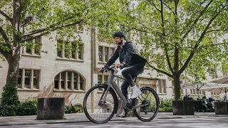 Ein Mann fährt mit einem E-Bike eine Straße entlang. Er trägt einen schwarzen Helm und eine graue Jacke. Im Hintergrund sind Bäume mit grünen Blättern zu sehen.