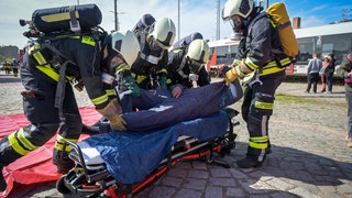 Feuerwehrleute legen bei einer Übung für einen Rettungsübungseinsatz eine Person auf eine Trage. Sie tragen Helme und Sauerstoffflaschen. Im Hintergrund sieht man blauen Himmel.