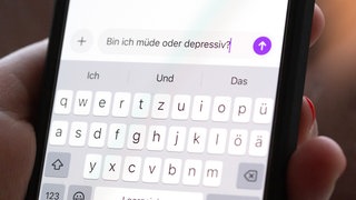 Eine Person hält ein Smartphone in der Hand, auf dem eine Suchanfrage mit der Frage "Bin ich müde oder depressiv?" in einem KI-Chatfenster zu sehen ist.