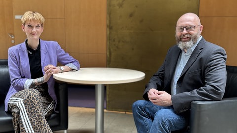 Queer-Beauftragte Sophie Koch und Korrespondent Dietrich Karl Mäurer sitzen in der Halle im ARD-Hauptstadtstudio in Sesseln an einem Tisch