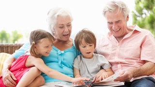 Oma und Opa schauen sich gemeinsam mit ihrer Enkeltochter und ihrem Enkelsohn ein Buch an. Symbolfoto
