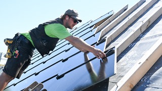 Ein Handwerker arbeitet an einem Photovoltaik Dach. Die neuartige PV Anlage besteht aus Photovoltaik Modulen in Form von Dachplatten. Es ist gutes Wetter, die Sonne scheint. Der Mann trägt eine Mütze und eine Sonnenbrille.