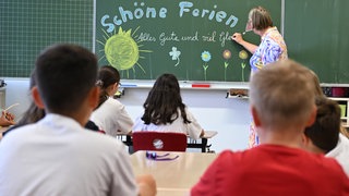 Lehrerin schreibt "Schöne Ferien" auf eine Tafel in einem Klassenzimmer voller Schüler.