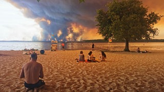 Strandbesucher sitzen am Strand von Moutchic in Lacanau im Südwesten Frankreichs, während Rauchwolken von einem Waldbrand in den Himmel aufsteigen.