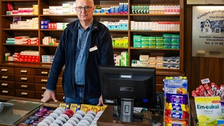 Hans-Günter Lund steht in seiner Apotheke. Hinter ihm sind die Regal voll mit Medikamenten. Er steht hinter dem Tresen, vor ihm ein Computerbildschirm.