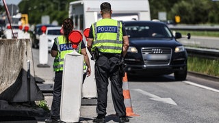 Polizisten der Bundespolizei kontrollieren Verkehrsteilnehmer am Grenzübergang Elten auf der A3 bei der Einreise nach Deutschland. Sie stehen mit dem Rücken zur Kamera und tragen Warnwesten mit der Aufschrift "Polizei". Vor ihnen bildet sich ein Stau.