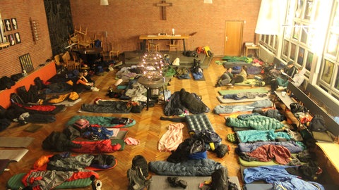 In einem Altarraum liegen unzählige Isomatten und Schlafsäcke auf dem Boden. Man sieht an der Wand ein Kreuz.