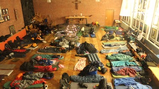 In einem Altarraum liegen unzählige Isomatten und Schlafsäcke auf dem Boden. Man sieht an der Wand ein Kreuz.