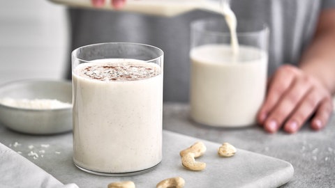 Ein Mann füllt ein großes Glas mit einer Flüssigkeit, die wie Milch aussieht. Da Nüsse auf dem Tisch liegen, könnte es aber auch ein Milch-Ersatzprodukt sein.