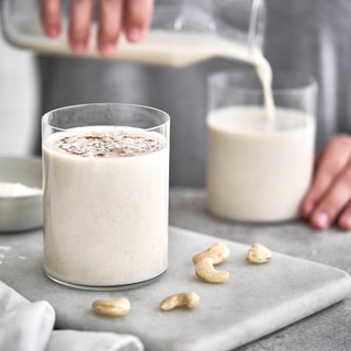Ein Mann füllt ein großes Glas mit einer Flüssigkeit, die wie Milch aussieht. Da Nüsse auf dem Tisch liegen, könnte es aber auch ein Milch-Ersatzprodukt sein.