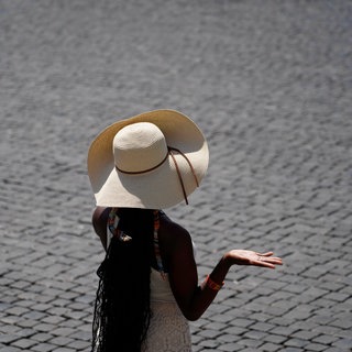 Eine Frau versucht sich mit einem Hut vor der Sonne zu Schützen. Sie ist auf einem Pflasterstein-Platz. Solche Plätze sind der Grund, warum viele Städte in Rheinland-Pfalz beim Hitze-Check der Deutschen Umwelthilfe schlecht abschneiden.