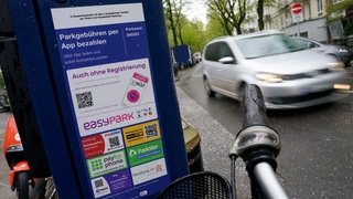 Aufkleber mit Logos von verschiedenen Handy-Park-Apps kleben auf einem Parkautomaten. DUH fordert deutlich höhere Gebühren: Parkschein darf nicht günstiger sein als ein Ticket für Bus und Bahn.