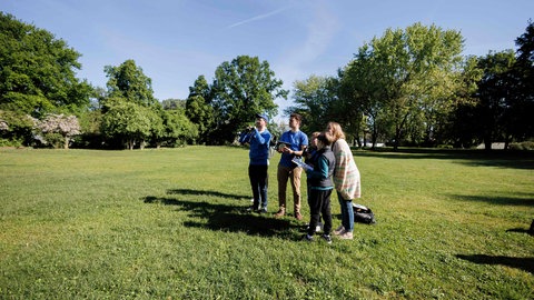 Mitglieder des Naturschutzbundes Deutschland (NABU) stehen auf einer Wiese und zählen mithilfe von Ferngläsern und Gehör das verschiedene Zwitschern von Vögeln zu Beginn der "Stunde der Gartenvögel" im Jahr 2024.