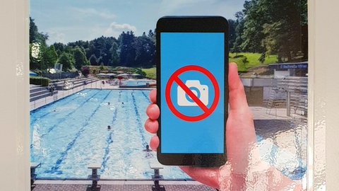 Ein Schwimmbecken im Freibad. Im Vordergrund eine Hand die ein Handy hält. Darauf ist ein durchgestrichenes Kamerasymbol abgebildet. Fotografieren ist oft im Freibad nicht erlaubt.