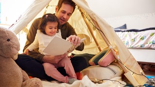 Ein Vater liest seiner Tochter aus einem Buch vor. (Symbolbild)