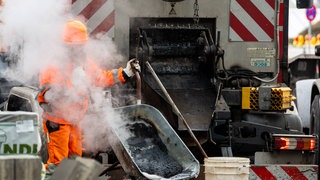 Dichter Dampf steigt auf, während ein Arbeiter heißen Asphalt aus der Maschine in die Schubkarre füllt. Auch die Bauwirtschaft leidet unter den Folgen des Irankriegs. (Symbolbild)