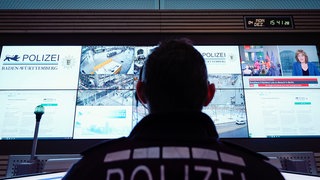 Ein Polizist sitzt in einem Lagezentrum vor einer Wand mit Bildschirmen, auf dem Bilder von Überwachungskameras zu sehen sind. Die Polizei in Baden-Württemberg soll eine Überwachungssoftware des umstrittenen Palantir-Konzerns einsetzen dürfen. Doch innerhalb der Grünen gibt es Kritik. (Symbolfoto)