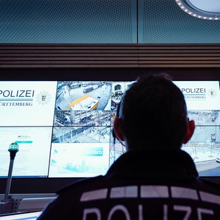 Ein Polizist sitzt in einem Lagezentrum vor einer Wand mit Bildschirmen, auf dem Bilder von Überwachungskameras zu sehen sind. Die Polizei in Baden-Württemberg soll eine Überwachungssoftware des umstrittenen Palantir-Konzerns einsetzen dürfen. Doch innerhalb der Grünen gibt es Kritik. (Symbolfoto)