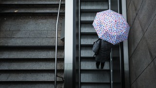 Eine Frau fährt am Dienstag mit ihrem Regenschirm auf der Rolltreppe einer Stuttgarter U-Bahn-Station. Auch in den kommenden Tagen bleibt es regnerisch in Baden-Württemberg. 