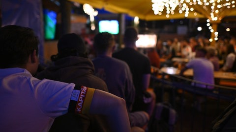 Gäste sitzen in einem Biergarten und sehen sich ein Spiel der WM 2024 an.