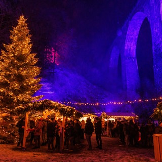 Menschen stehen zwischen Verkaufsständen auf dem Weihnachtsmarkt in der Ravennaschlucht, der unter einem Aquädukt stattfindet. 