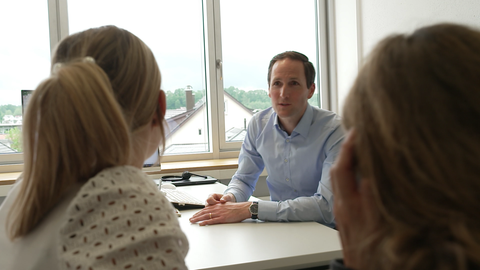Berater sitzt an einem Tisch zwei Frauen gegenüber, deren Hinterköpfe im Vordergrund zu sehen sind.
