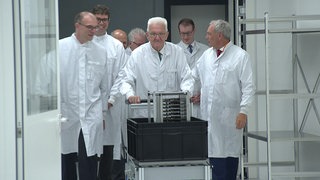 Ministerpräsident Winfried Kretschmann (Grüne) trifft sich mit Mitarbeitenden der Backnanger Firma Tesat in einem Unternehmensgebäude und trägt dabei wie sie einen weißen Laboranzug.